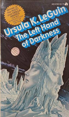 The cover of The Left Hand of Darkness by Ursula Le Guin shows an ice sculpture rising out of a frozen landscape. The sculpture has a woman’s face on side and a man’s face on the other side. The background shows a dark sky with a planet with rings around it. There is a sticker in the upper left corner declaring this book a Hugo and Nebula award winner.